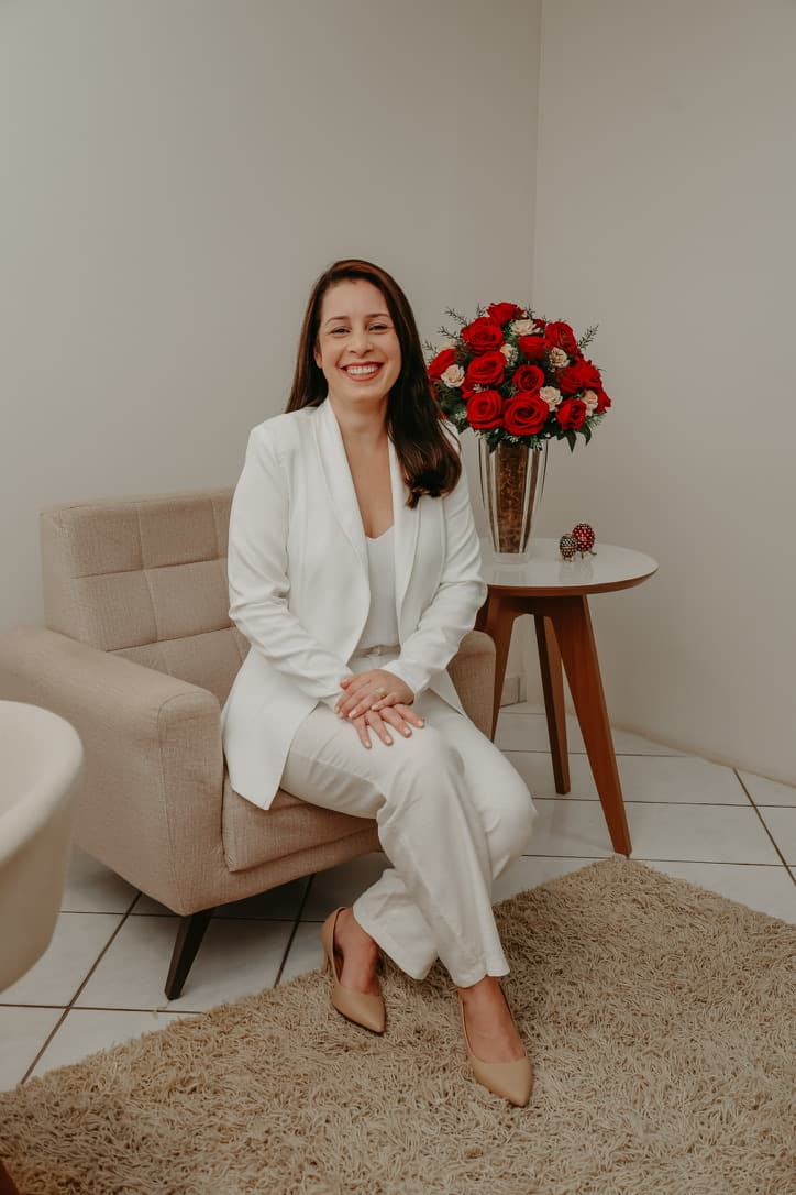 Viviane Cunha, psicóloga, sorrindo sentada em uma poltrona bege do consultório, ao lado de um arranjo de rosas vermelhas